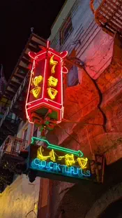 A neon sign in front of the Li-Po Lounge in San Francisco at night. A red and yellow neon sign in the shape of a Chinese lantern says “Li Po” in faux-Chinese lettering. Below it a yellow and blue sign says “Li-Po Cocktails”, with an arrow pointing toward the entrance.